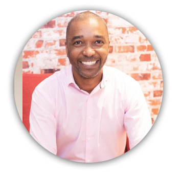 imagem circular. Alexandre é um homem negro, calvo. Está sorrindo e usa uma blusa social rosa. Ele está num ambiente bem iluminado.