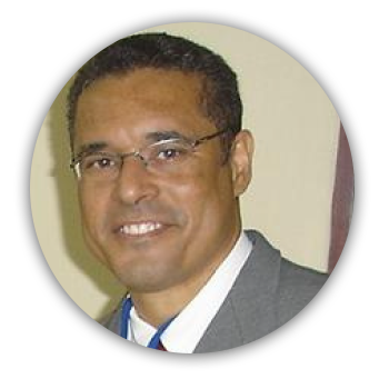 imagem circular.Celso Eduardo é um homem negro, ele está sorrindo. Celso usa óculos e está vestido com terno e gravata.