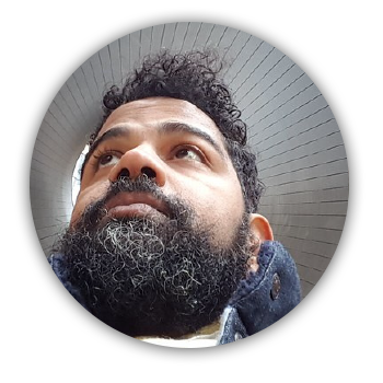 imagem circular. Paulo Rená é um homem negro. o Ãngulo da foto é de baixo para cima. Paulo Rená usa barba e olha para o canto direito. Acima dele dá para ver o teto cinza com faixas simétricas como se saíssem do centro do círculo.